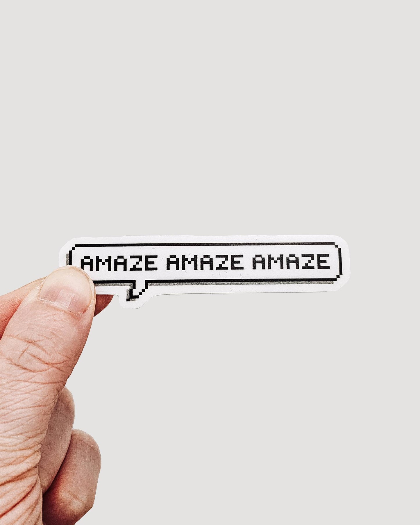 Hand holding a small sticker with 'AMAZE AMAZE AMAZE' text on a plain background