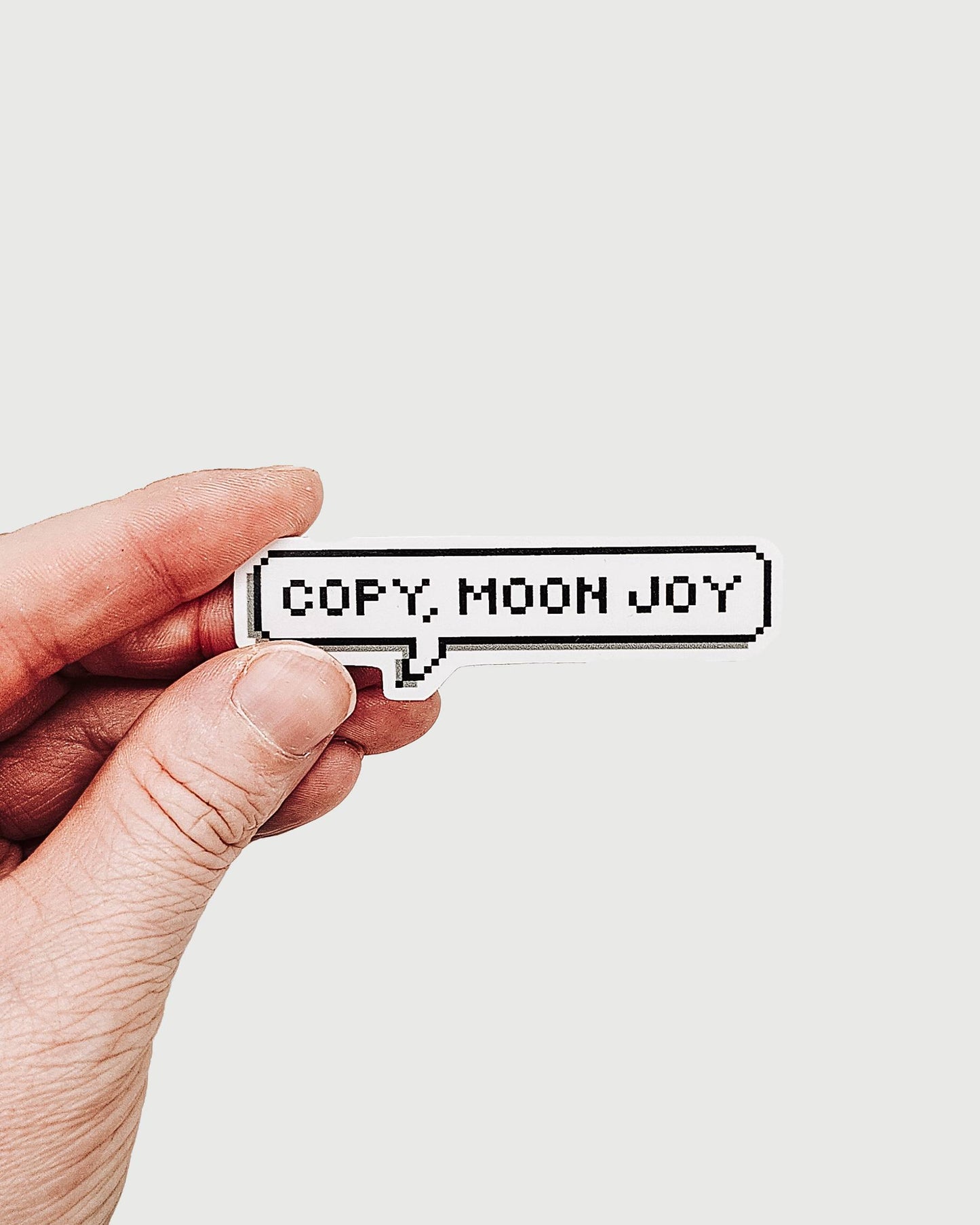 Hand holding a small rectangular sticker with 'COPY, MOON JOY' text on a light background